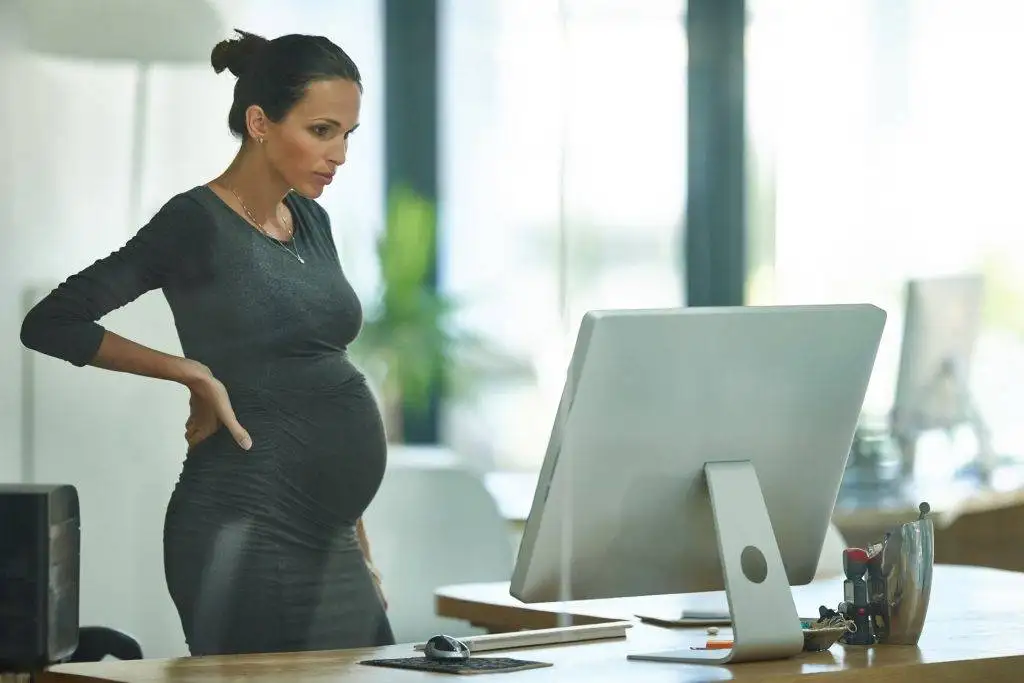 Pregnant woman standing at desk and working on desktop computer