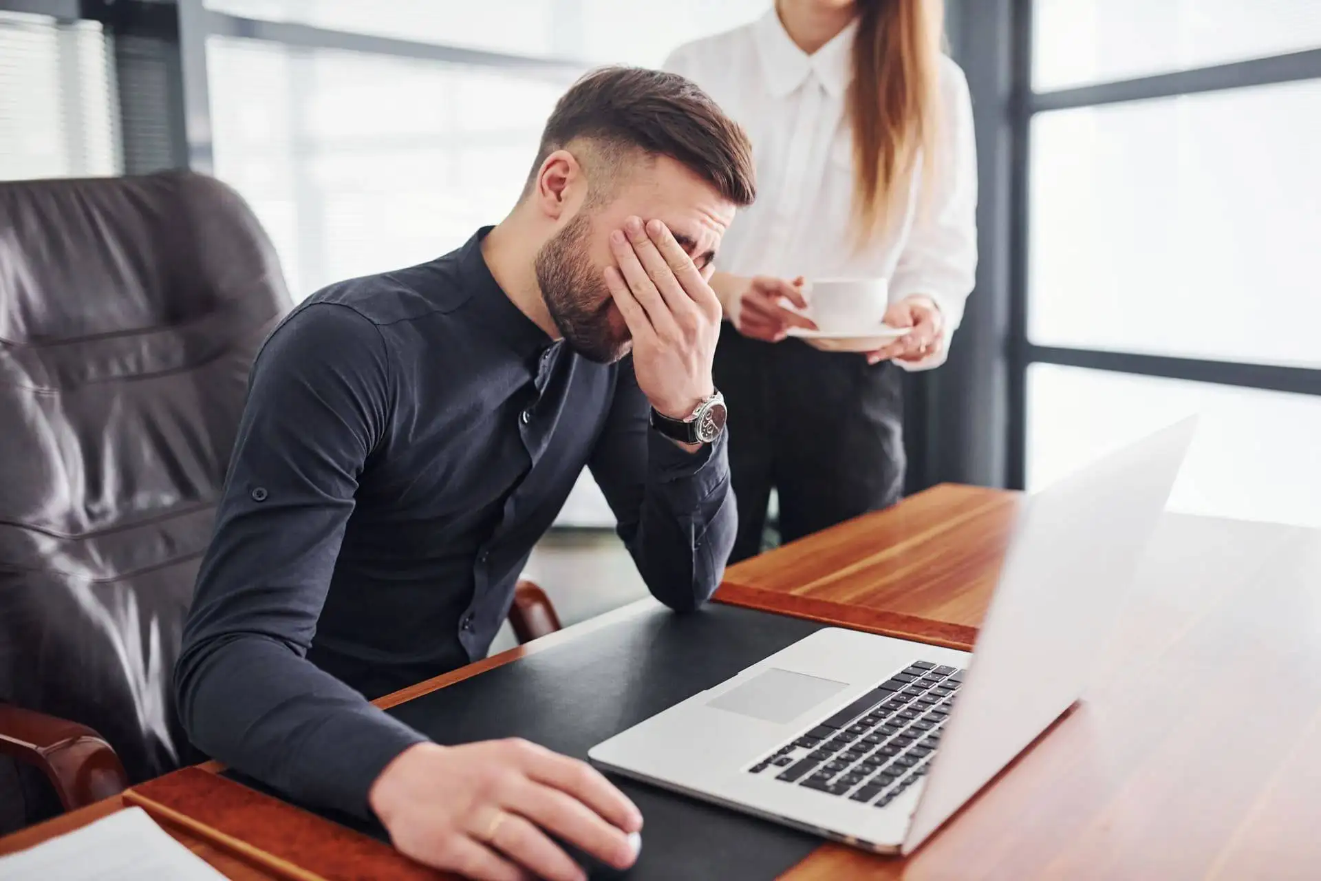 Stressed businessman sitting at a laptop with a woman standing nearby