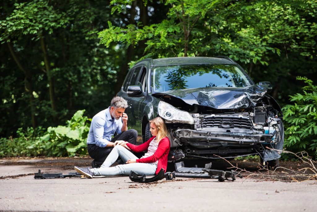 young woman sat next to a crashed car and a man on the phone